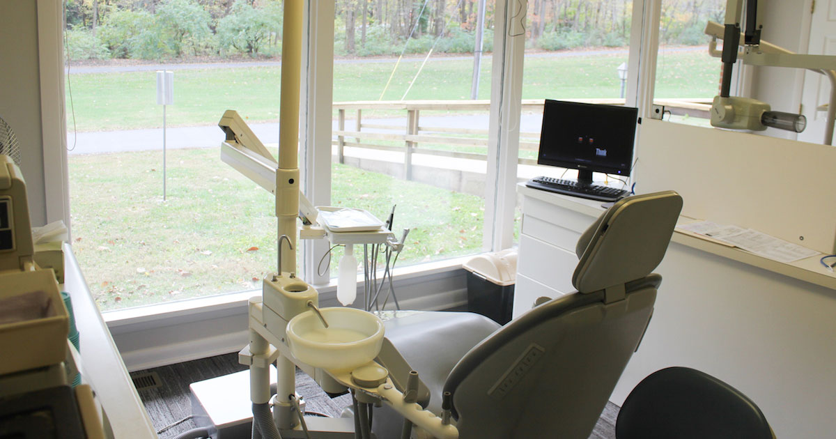 The image shows an interior view of a dental office with a large window, equipment, and a waiting area.