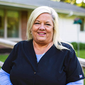The image shows a woman wearing a white lab coat, standing in front of a house with a lawn.