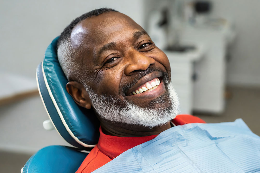 The image shows a man with a beard, wearing a red shirt, sitting in a dental chair with a smile on his face.