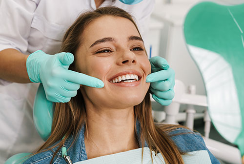 The image shows a woman sitting in a dental chair with her eyes closed, smiling broadly, while undergoing dental treatment  a dentist is adjusting her teeth with a smile on their face.