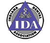 The image is a logo featuring the acronym  IDA  in large, bold letters, with the word  Association  directly below it. Above the acronym, there is an emblem that includes a stylized depiction of a dental implant and a circular design resembling a shield or badge. The logo has a central, rectangular shape with rounded corners, set against a background that transitions from white at the top to a darker shade towards the bottom.