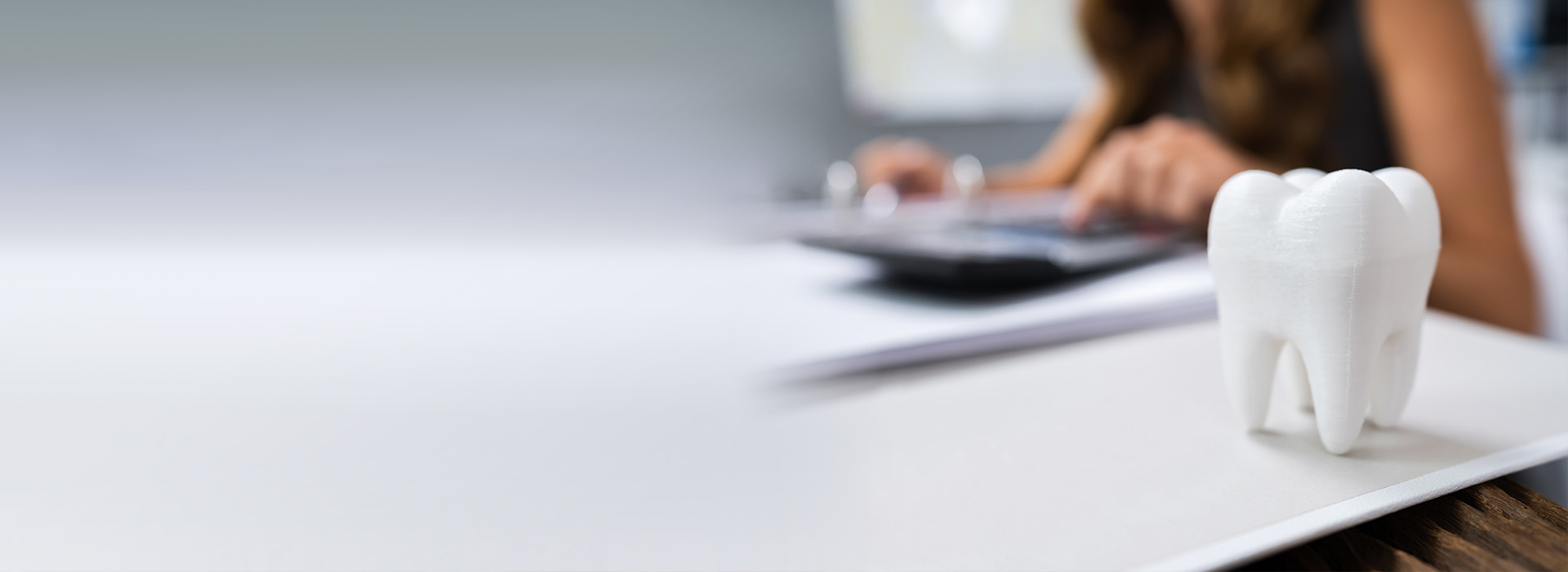 The image shows a person sitting at a desk with a computer, working on a document. There is a model of a tooth in the foreground, and the background features an office setting with a whiteboard and other office items.