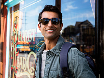 The image is a photograph of a man standing outdoors, wearing sunglasses and a backpack. He has a beard and mustache, and is dressed in casual attire with a jacket, t-shirt, and jeans. The background includes a shop window with the name Sweet Treats visible, suggesting an urban setting.