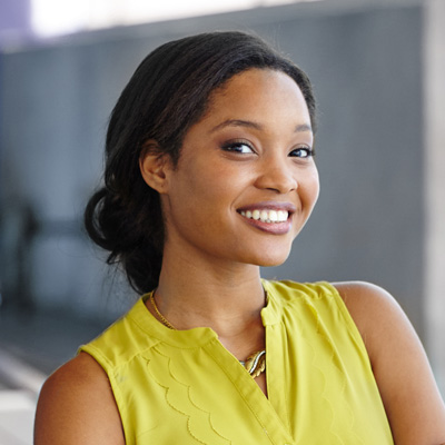 A woman with a big smile, wearing a yellow blouse and standing against a wall.