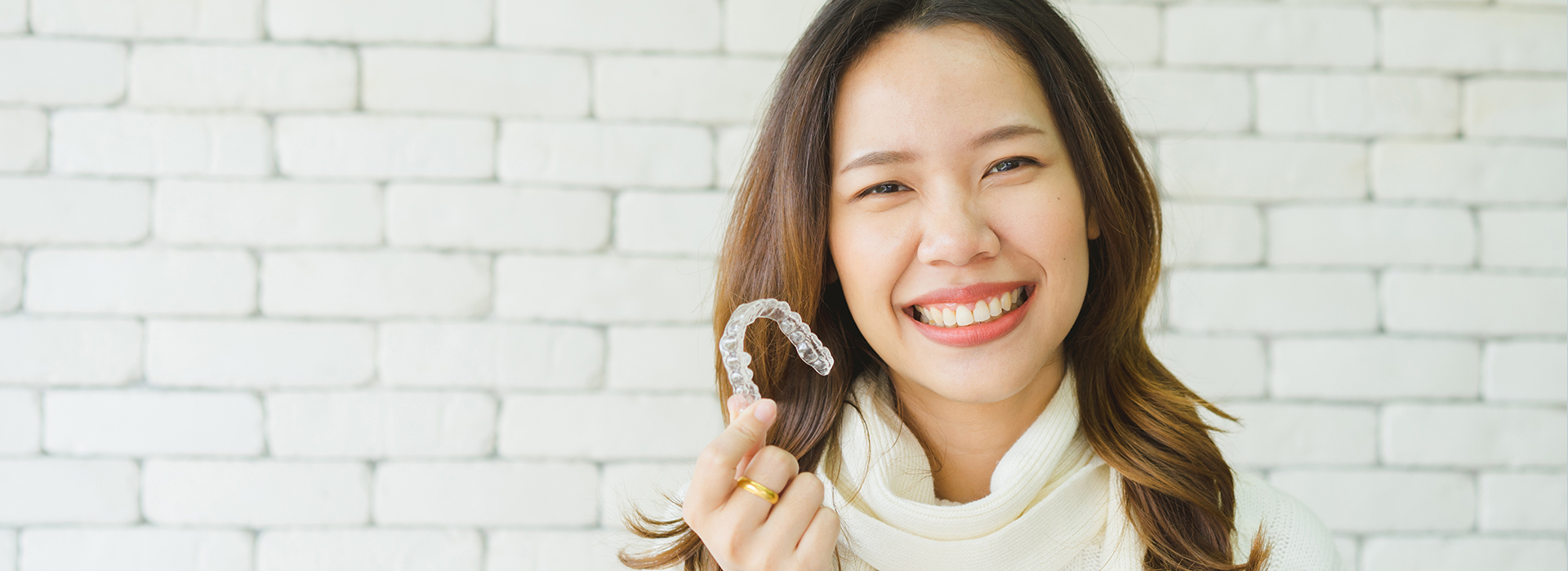 A woman with a smile is holding a ring in her hand, set against a brick wall background.