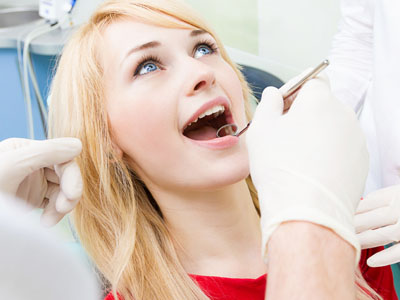 The image shows a woman sitting in a dental chair, receiving treatment from a dentist who is holding a dental drill and wearing protective eyewear.